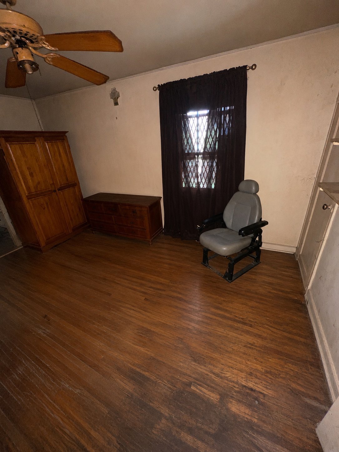 Sparsely furnished room with wooden floor, wooden wardrobe, black curtained window, ceiling fan, and gray gaming chair