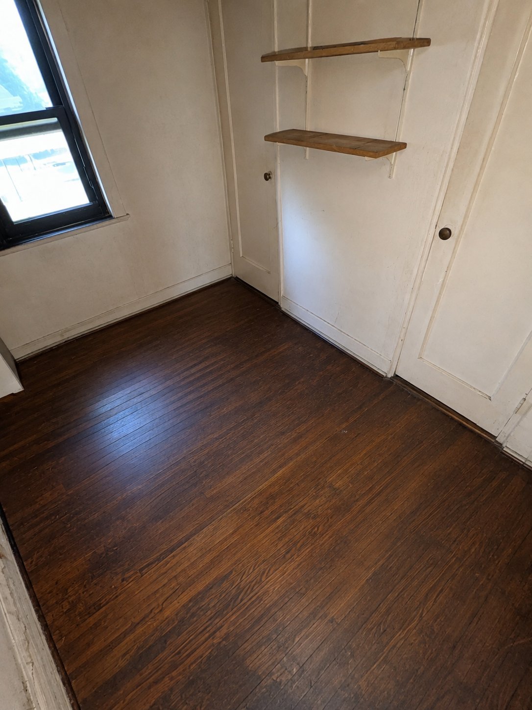 Empty room with dark hardwood flooring, white walls, window, and two wooden shelves mounted on wall