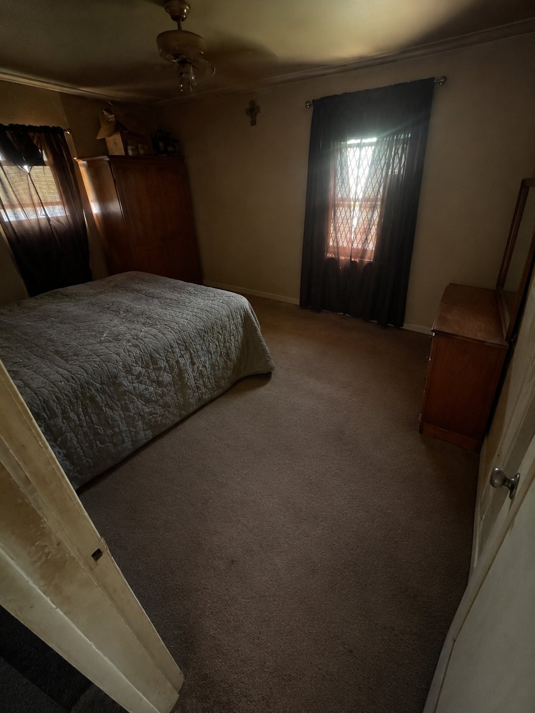 Spacious bedroom with a queen-size bed, dark curtains on two windows, wooden nightstands, ceiling fan, and beige walls.
