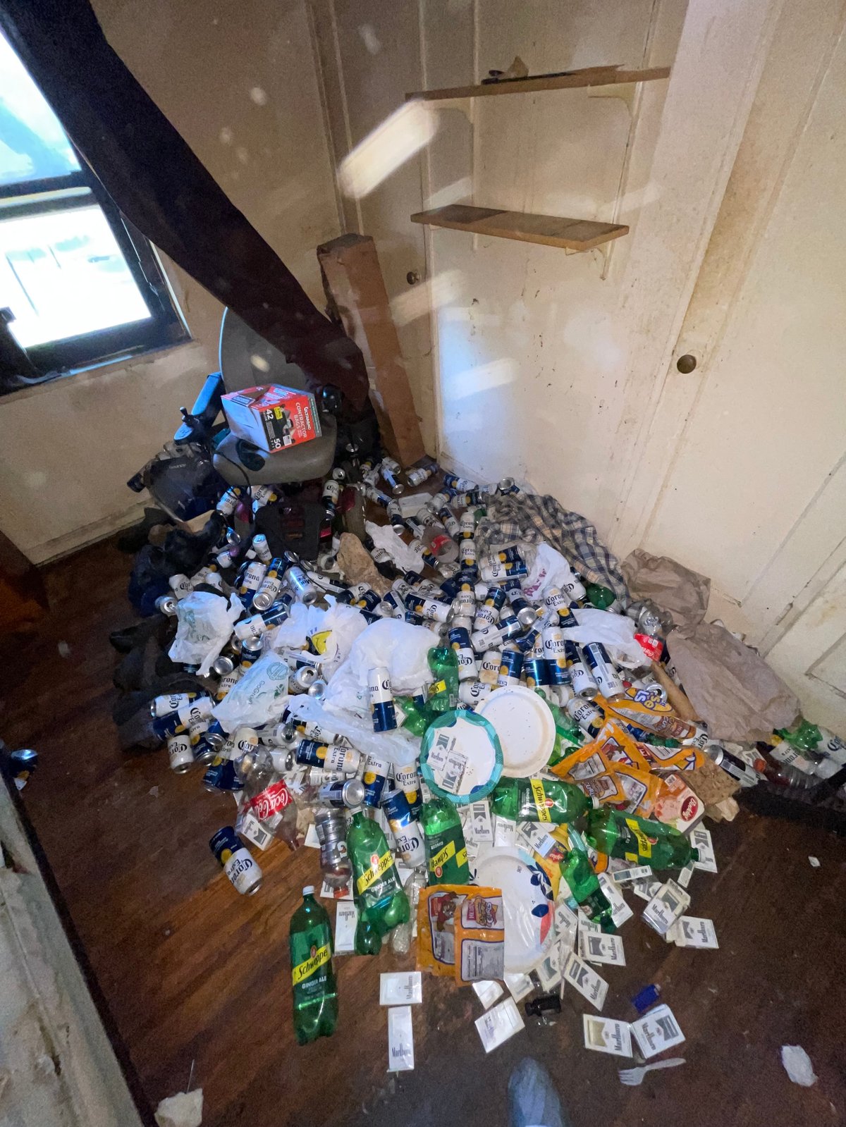 Large pile of mixed trash and discarded items scattered on a wooden floor in an empty room with wooden shelves.