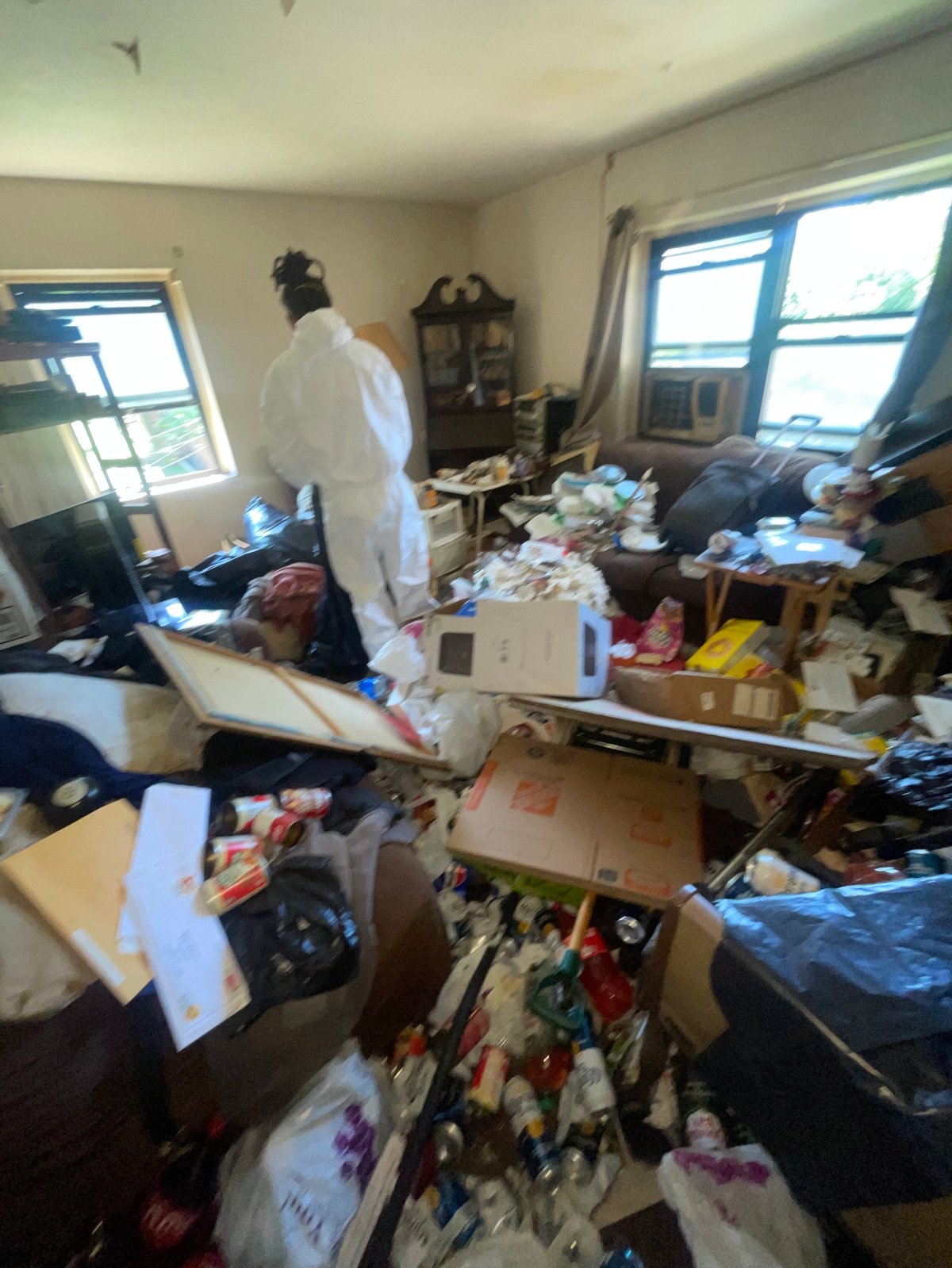 Cluttered bedroom with person in white protective suit standing amid piles of boxes, clothing, and items scattered across floor and furniture