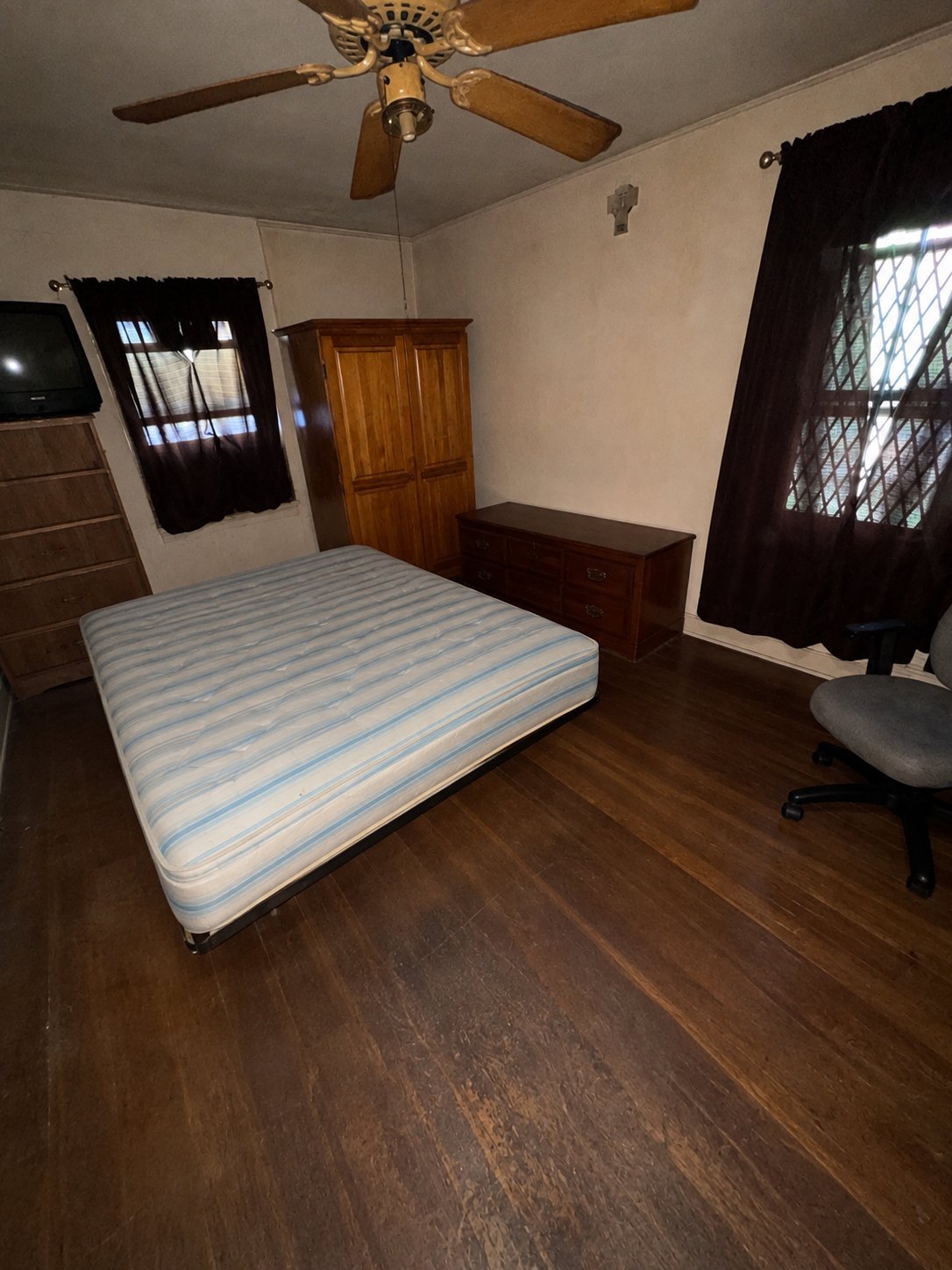 Bedroom interior with striped mattress on floor, ceiling fan, wooden furniture, and black curtains on windows