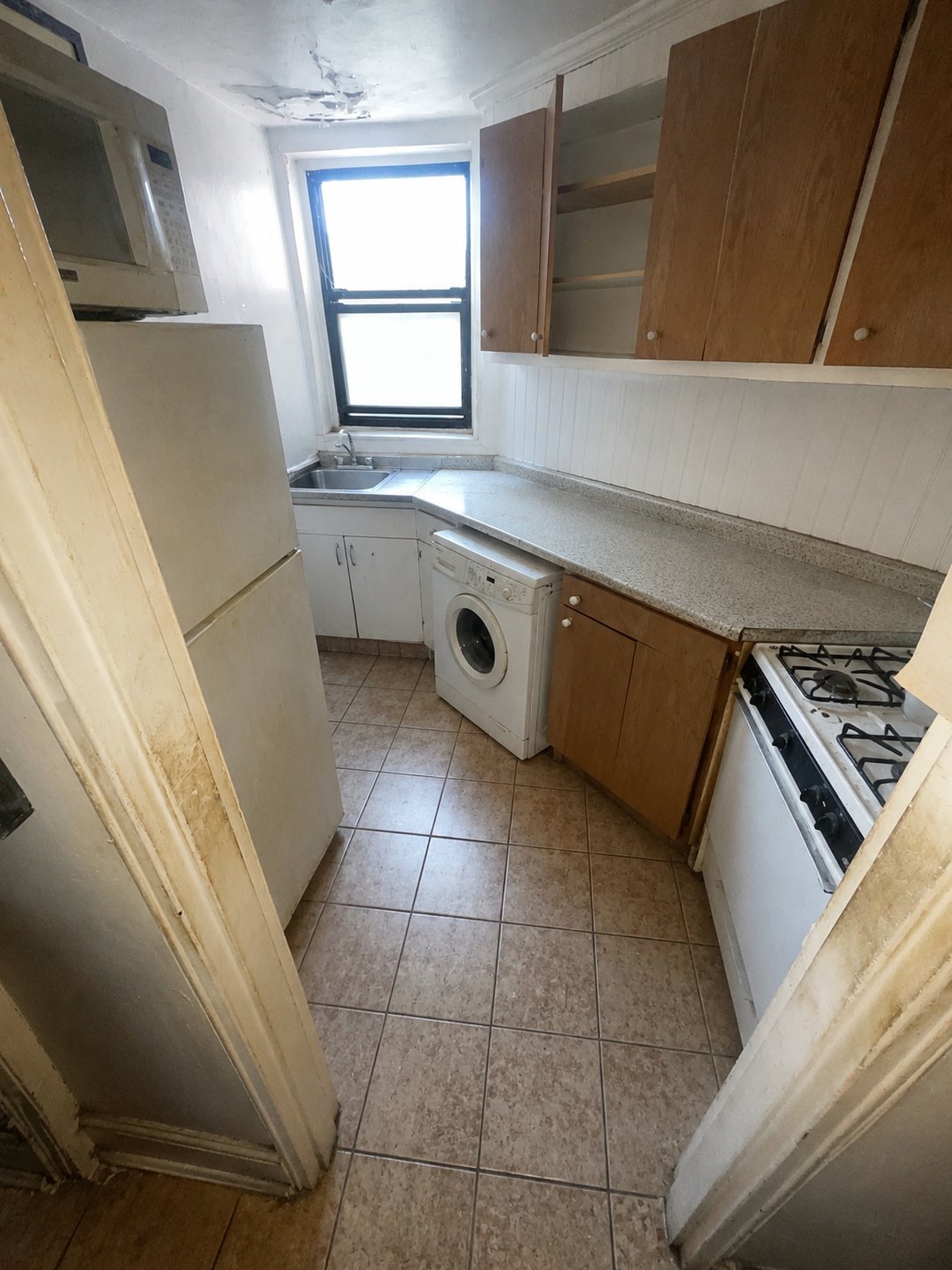 Narrow galley kitchen with white appliances, beige countertop, brown cabinetry, washing machine, tiled floor, and window with blue trim