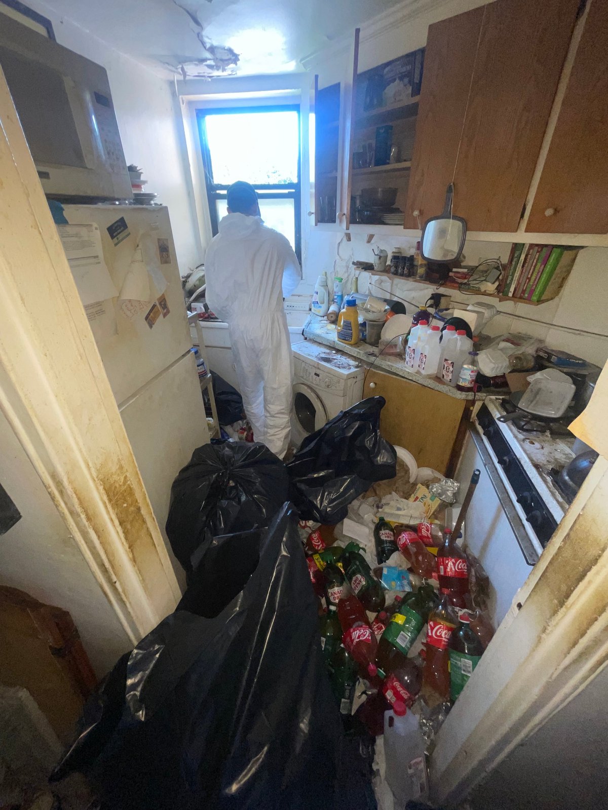 Person in white protective suit cleaning cluttered kitchen with trash bags, bottles, and recycling materials visible