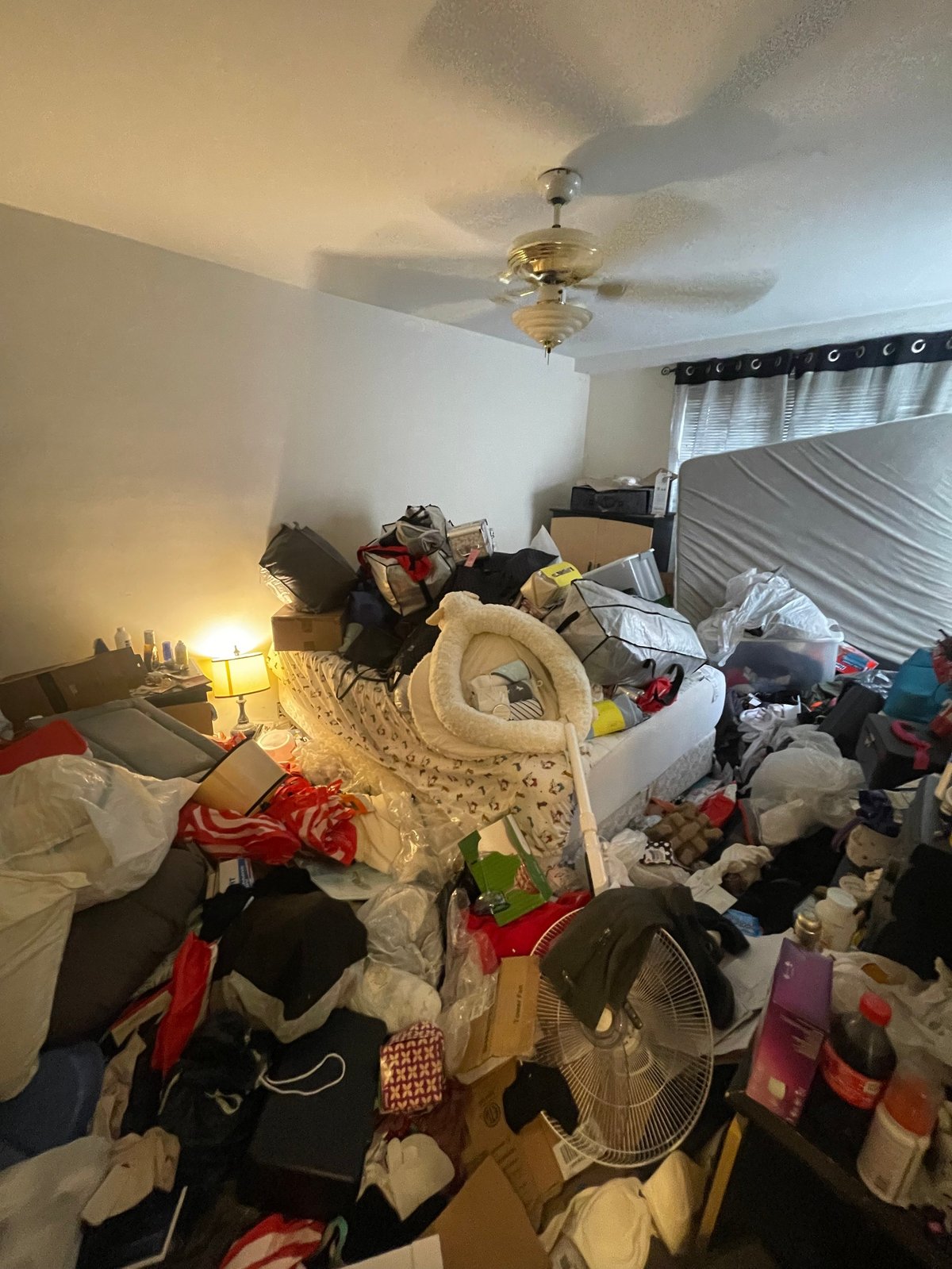 Cluttered bedroom with piles of clothes and belongings scattered across the floor and furniture