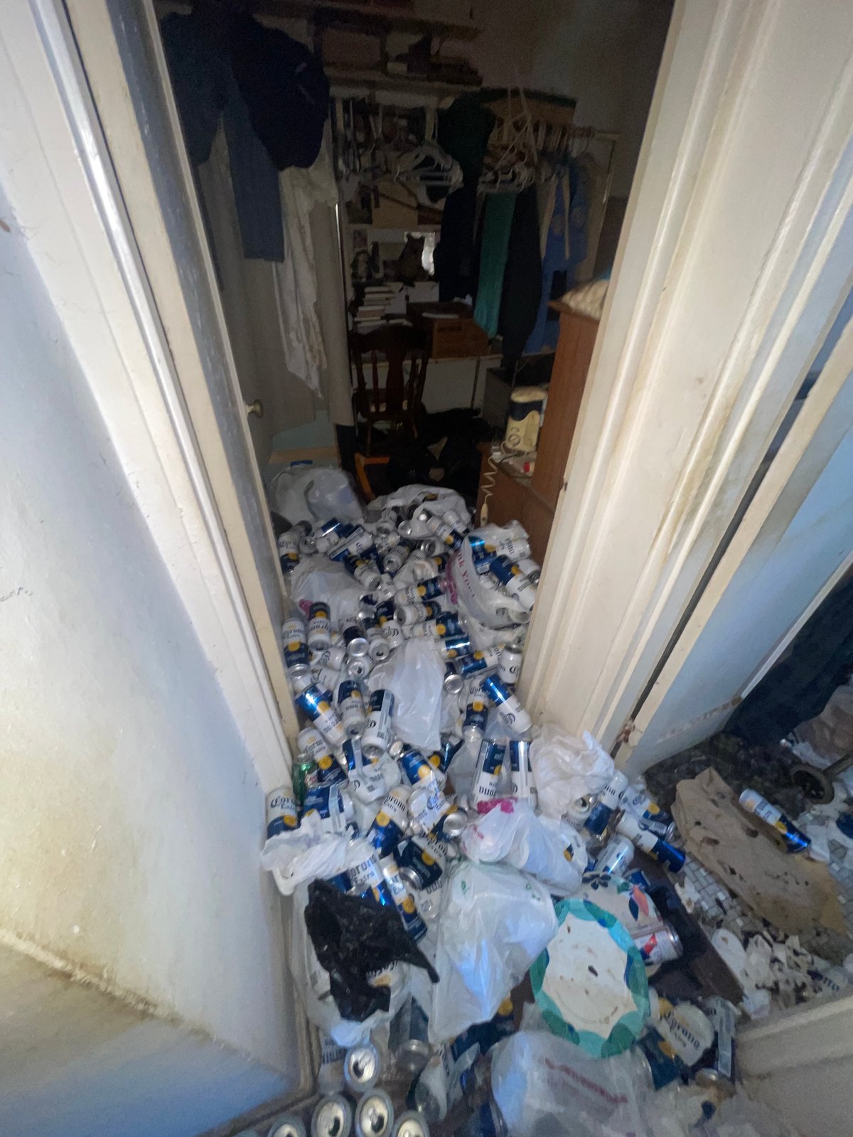 Cluttered closet filled with piles of plastic bottles, clothes, and miscellaneous items overflowing onto the floor through open double doors