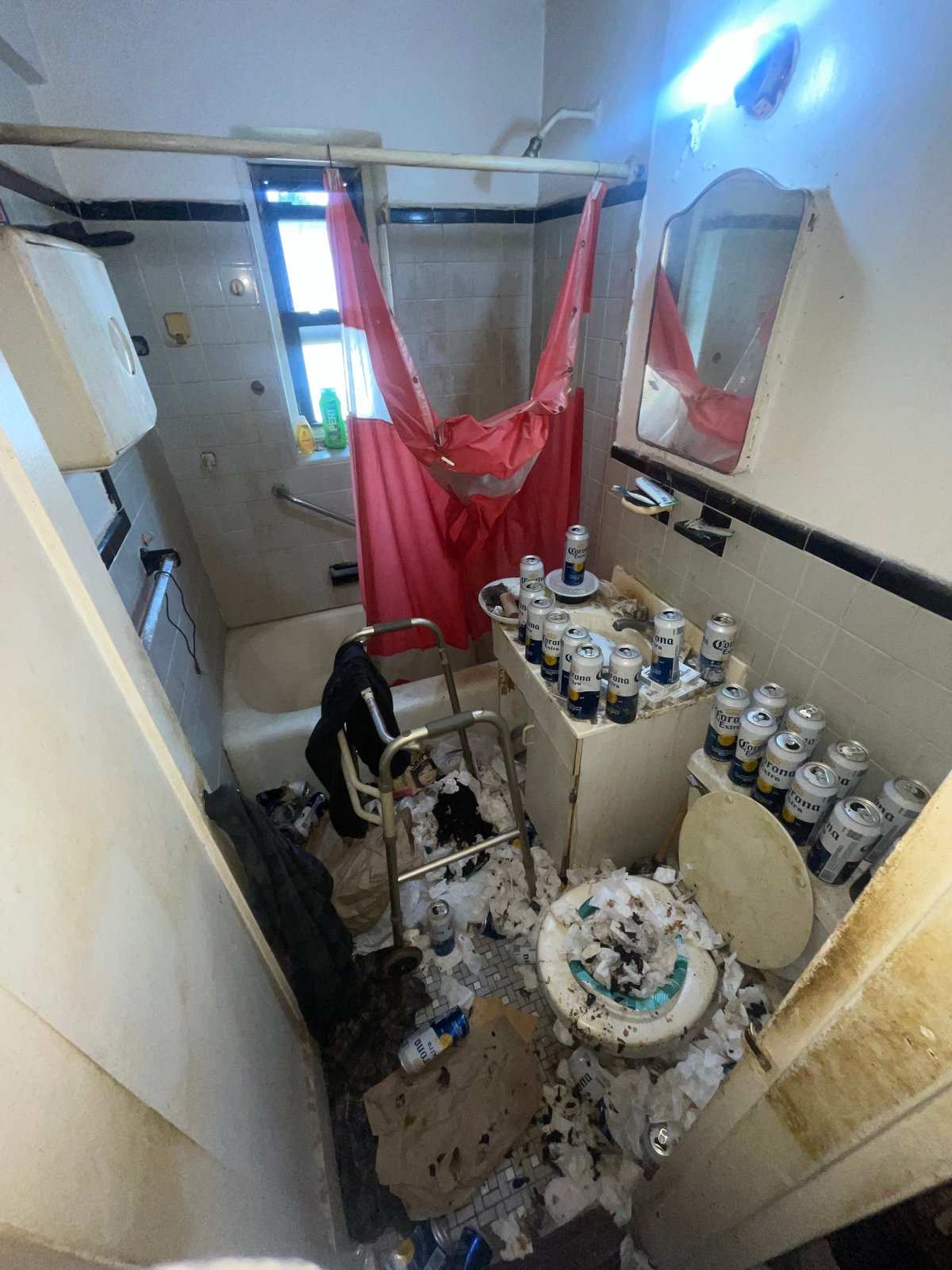 Cluttered bathroom with red shower curtain, jars and containers scattered on surfaces, chair, and items on floor