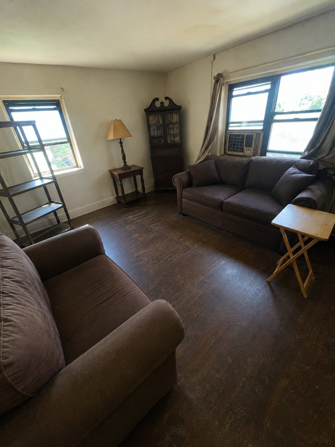 Bright living room with brown leather furniture, dark wood floors, windows with blue frames, and a wooden side table with lamp