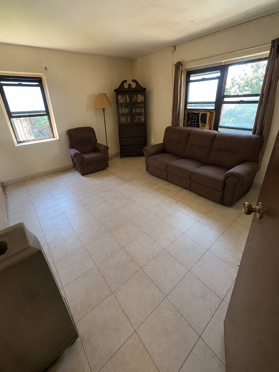Spacious living room with brown recliner and sofa, beige walls, tiled floor, bookshelf, floor lamp, and large windows overlooking outdoor area
