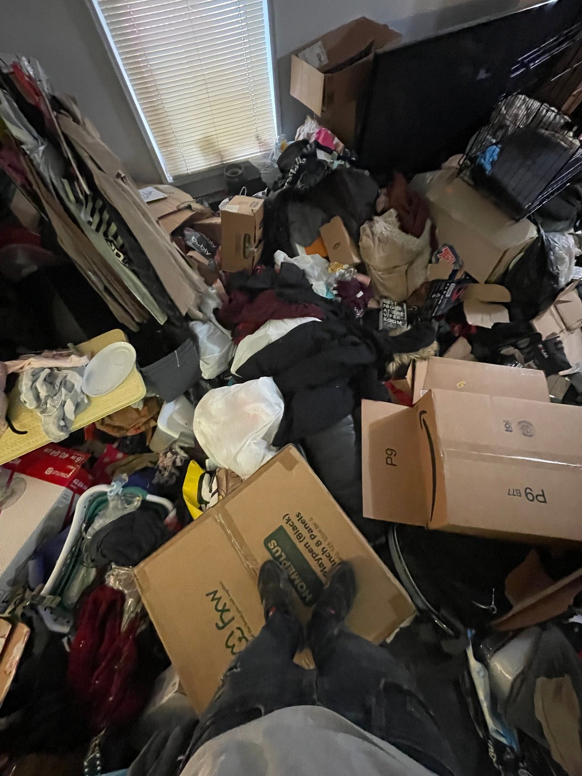 Cluttered room filled with cardboard boxes, black plastic bags, clothes, and miscellaneous items scattered across the floor